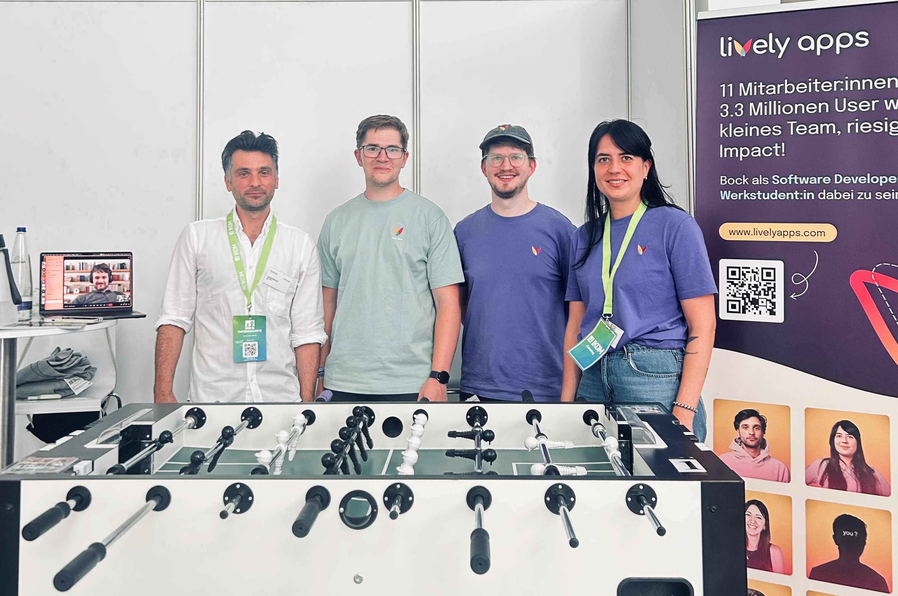 Four team members from Lively Apps standing behind a football table at the IKOM 2025 career fair booth, with a company banner and laptop setup in the background.